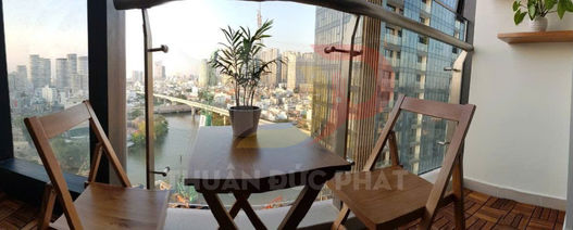 Bàn ghế gỗ ban công nhỏ với chậu cây xanh, lan can kính nhìn ra sông và các tòa nhà cao tầng