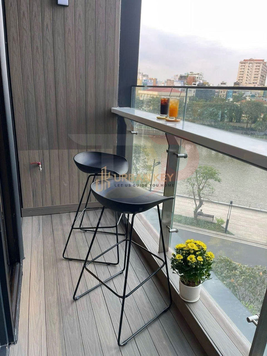 Ban công nhỏ với hai ghế bar chân sắt, bàn kính đặt hai ly nước và chậu hoa cúc vàng nhìn ra sông