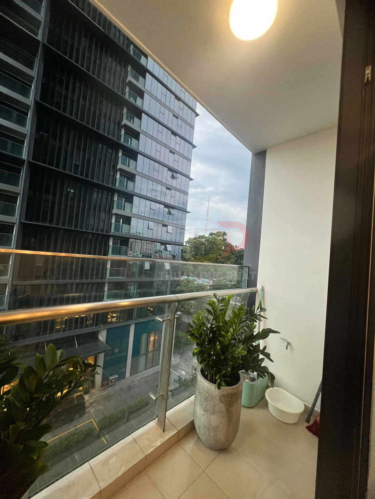 Ban công chung cư với lan can kính, chậu cây xanh trang trí và tòa nhà văn phòng hiện đại phía trước