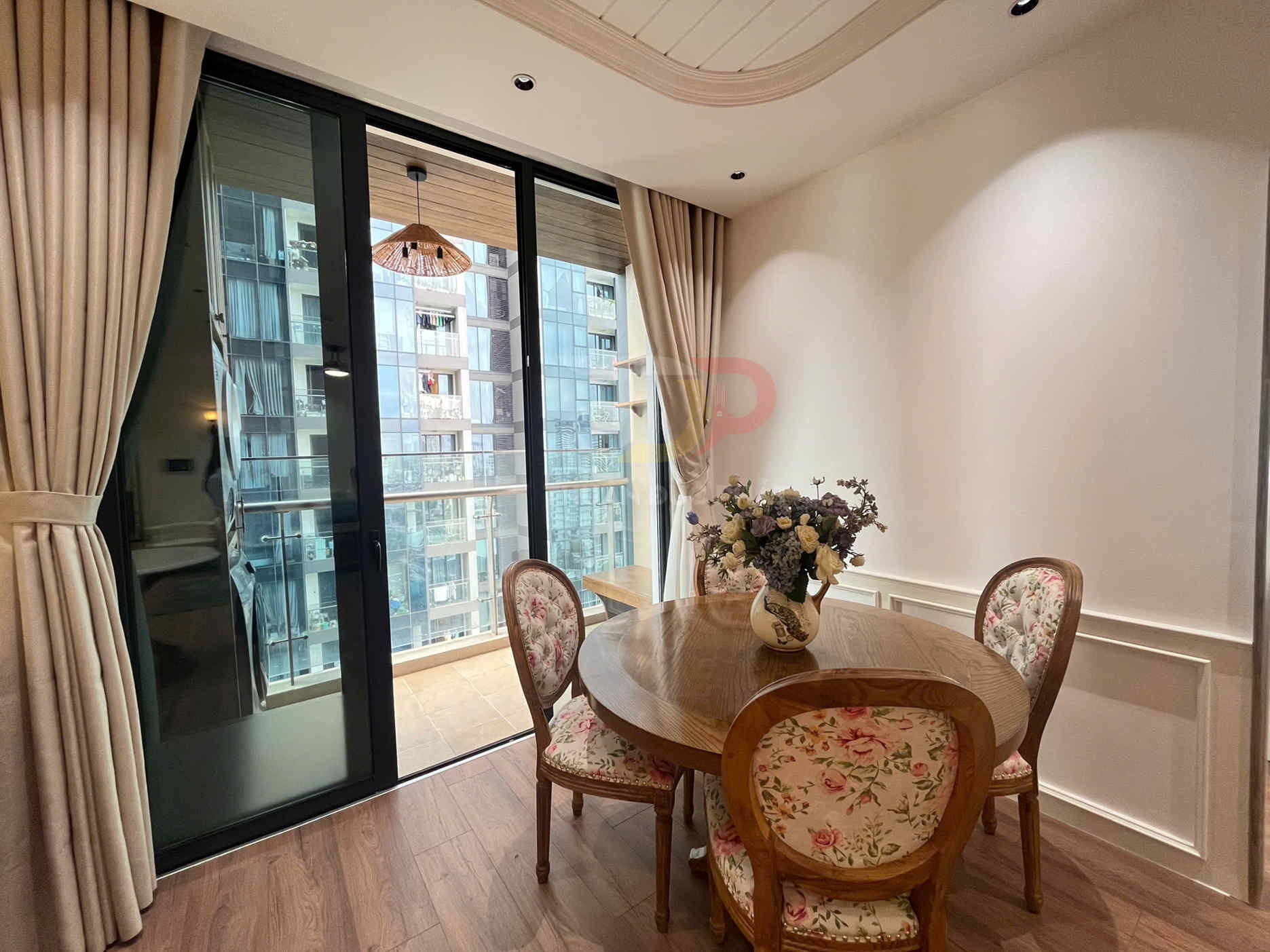 Bàn ăn gỗ tròn với ghế bọc nệm hoa và bình hoa trang trí cạnh cửa kính ban công căn hộ