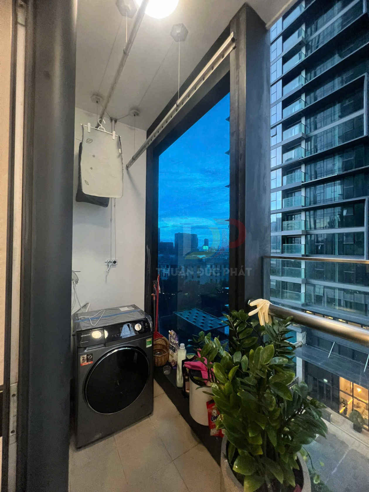 Ban công chung cư với máy giặt cửa trước màu đen và cây xanh cạnh cửa kính nhìn ra tòa nhà cao tầng