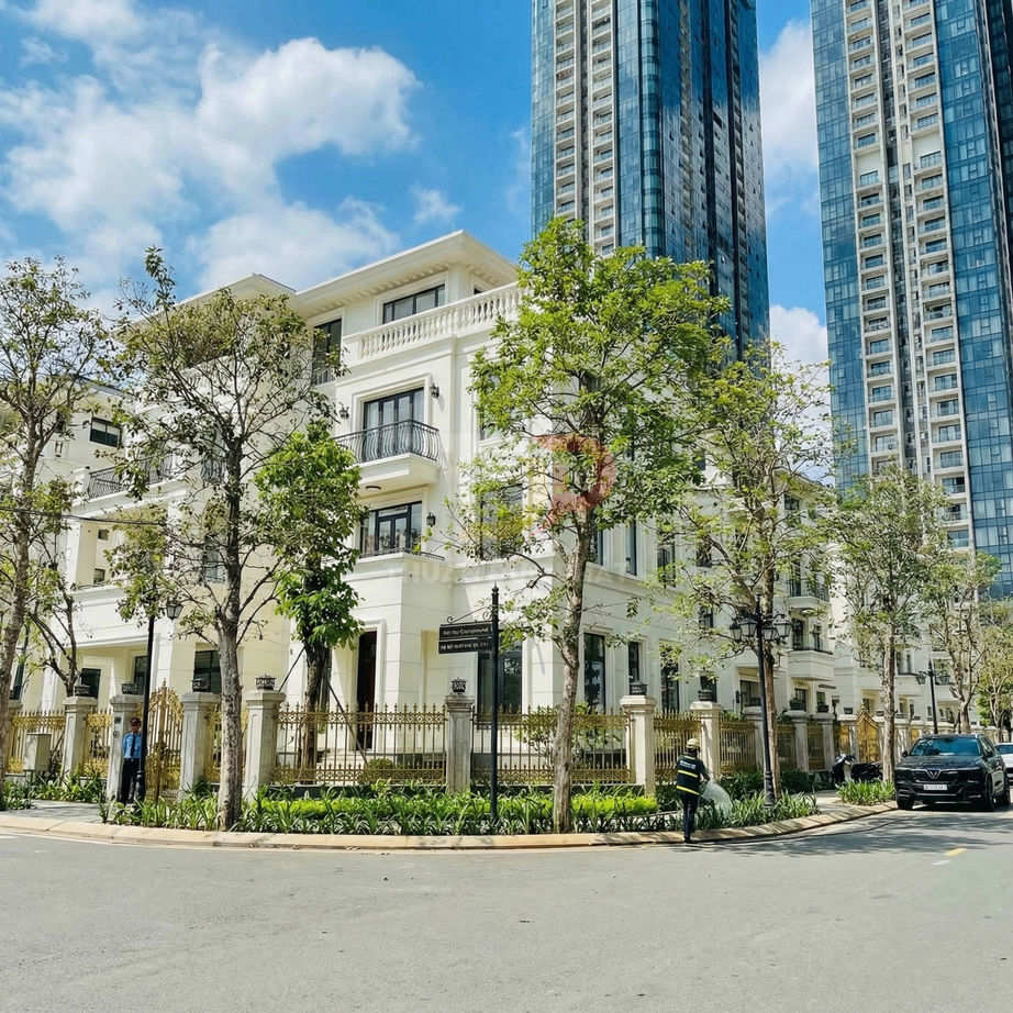Biệt thự trắng cao cấp góc phố với hàng rào sắt và cây xanh, phía sau là các tòa chung cư cao tầng