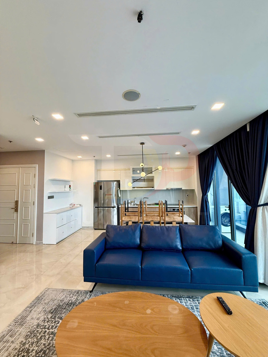 Sofa da màu xanh dương trong phòng khách căn hộ hiện đại cạnh bàn trà gỗ và bếp tích hợp tủ lạnh inox