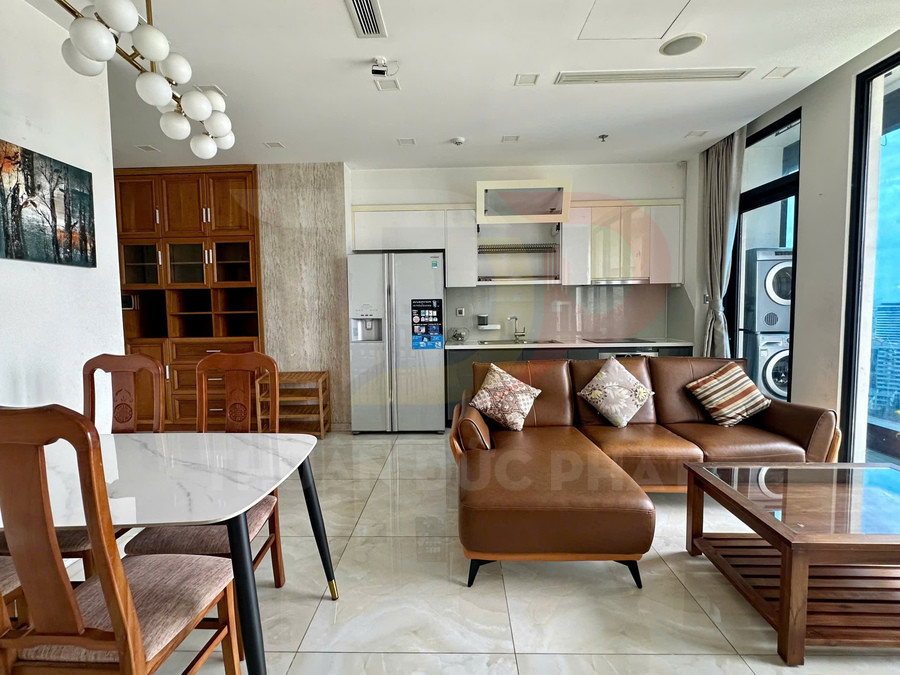 Phòng khách căn hộ với sofa da chữ L, bàn trà gỗ, bếp và tủ lạnh cạnh bàn ăn gỗ hiện đại