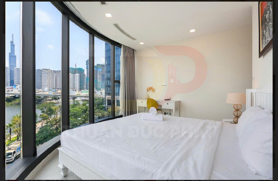 Phòng ngủ căn hộ cao cấp với giường đôi trắng cạnh cửa kính lớn nhìn ra sông và skyline thành phố