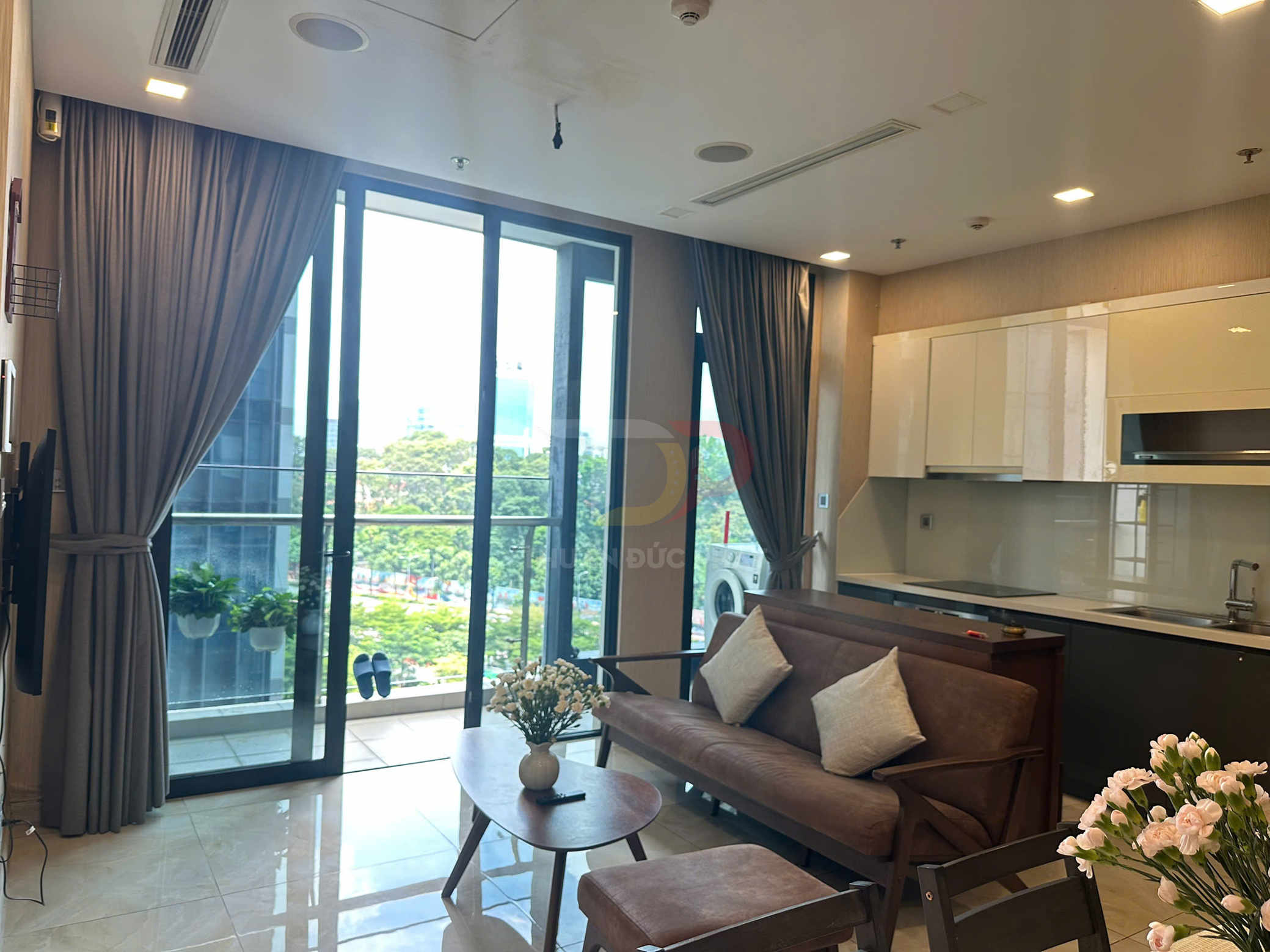 Phòng khách căn hộ hiện đại với sofa nâu, bàn trà gỗ, bếp liền kề và cửa kính lớn nhìn ra ban công