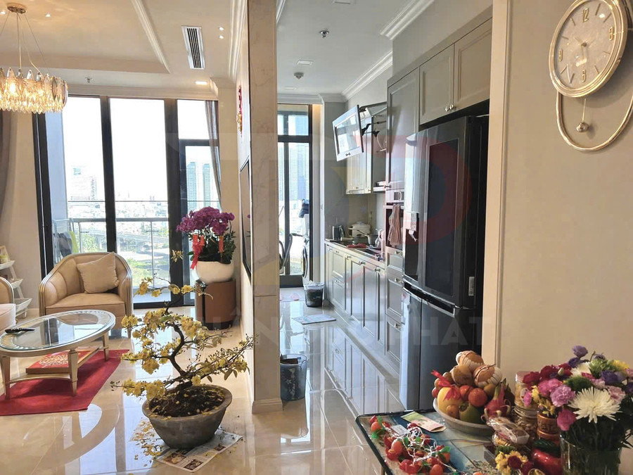 Phòng khách và bếp căn hộ hiện đại với cửa kính lớn, bàn ghế sofa, tủ bếp và tủ lạnh, trang trí hoa và mâm trái cây
