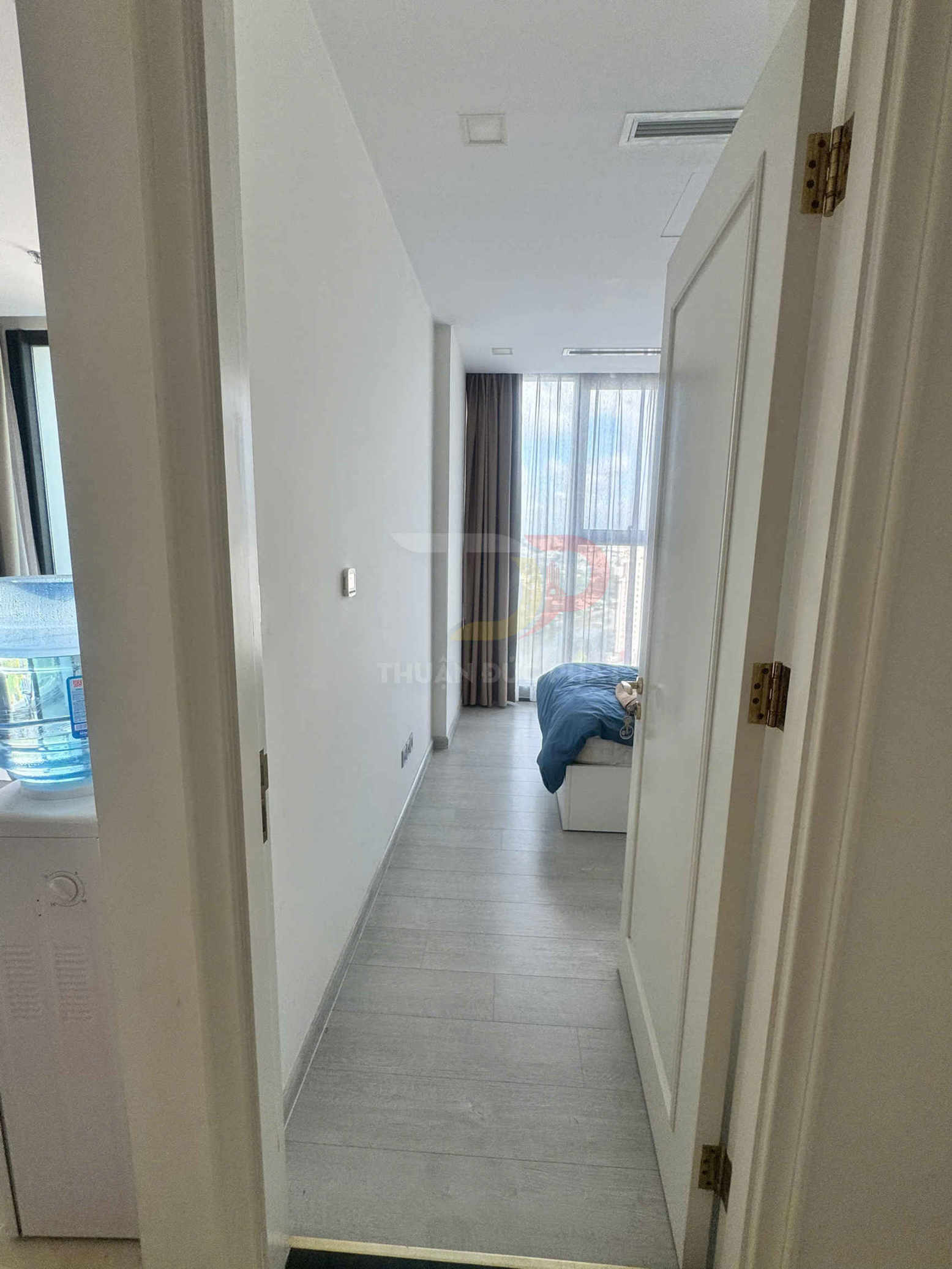 Lối vào phòng ngủ chung cư với giường phủ chăn xanh và cửa sổ lớn rèm xám sáng