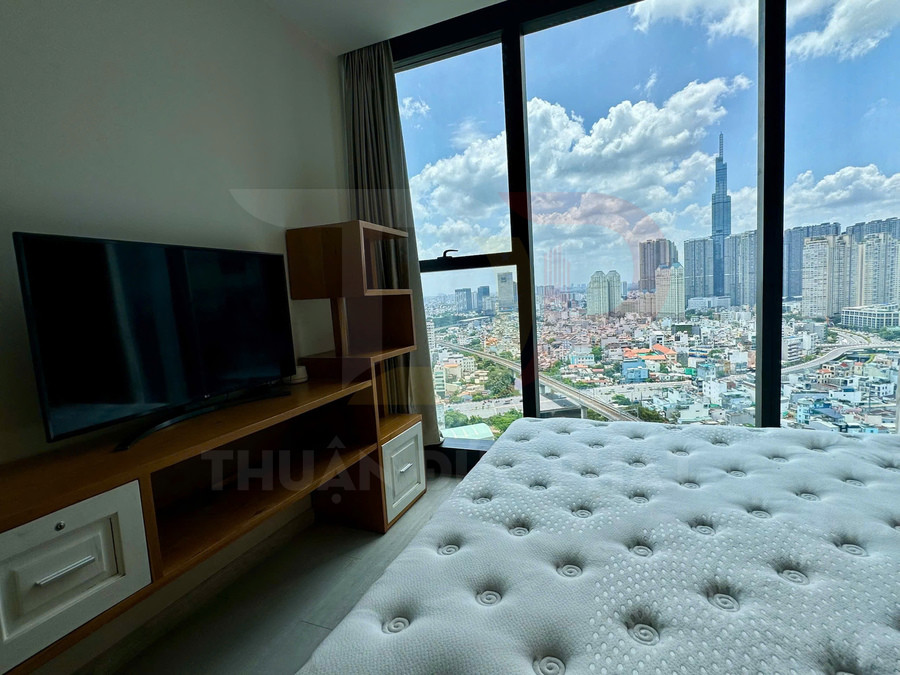 Phòng ngủ căn hộ với giường nệm trắng, tivi trên kệ gỗ và cửa sổ lớn nhìn ra thành phố