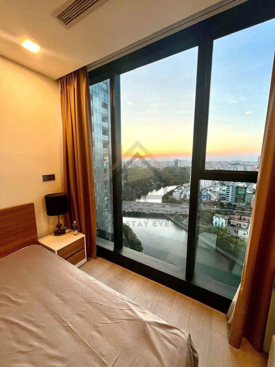 Phòng ngủ chung cư với giường đơn, cửa sổ kính lớn nhìn ra sông và thành phố lúc hoàng hôn