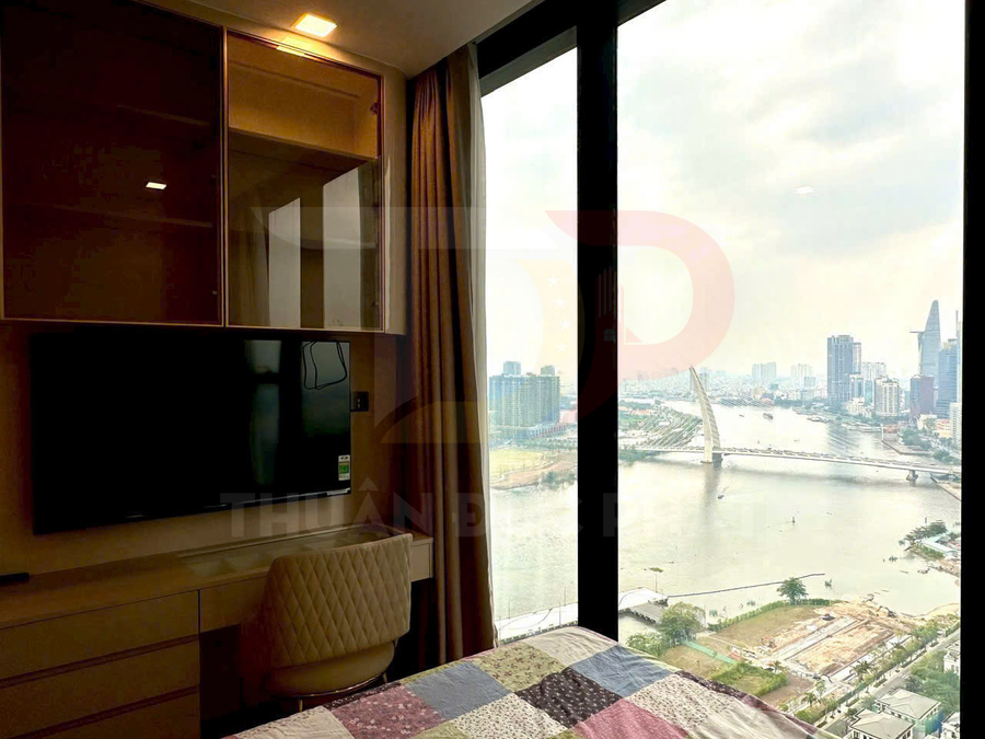 Phòng ngủ chung cư với bàn làm việc, tivi treo tường và cửa kính lớn nhìn ra sông và skyline thành phố