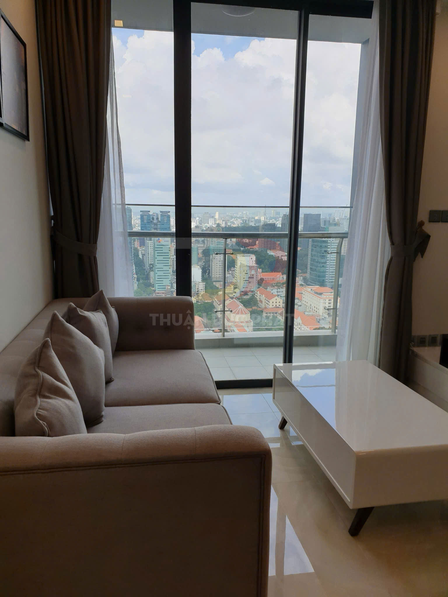 Sofa góc vải màu nâu nhạt với bàn trà trắng đặt cạnh cửa kính lớn nhìn ra thành phố