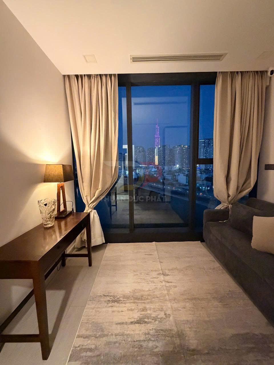 Phòng khách chung cư với sofa, bàn gỗ, đèn bàn và cửa kính lớn nhìn ra thành phố ban đêm