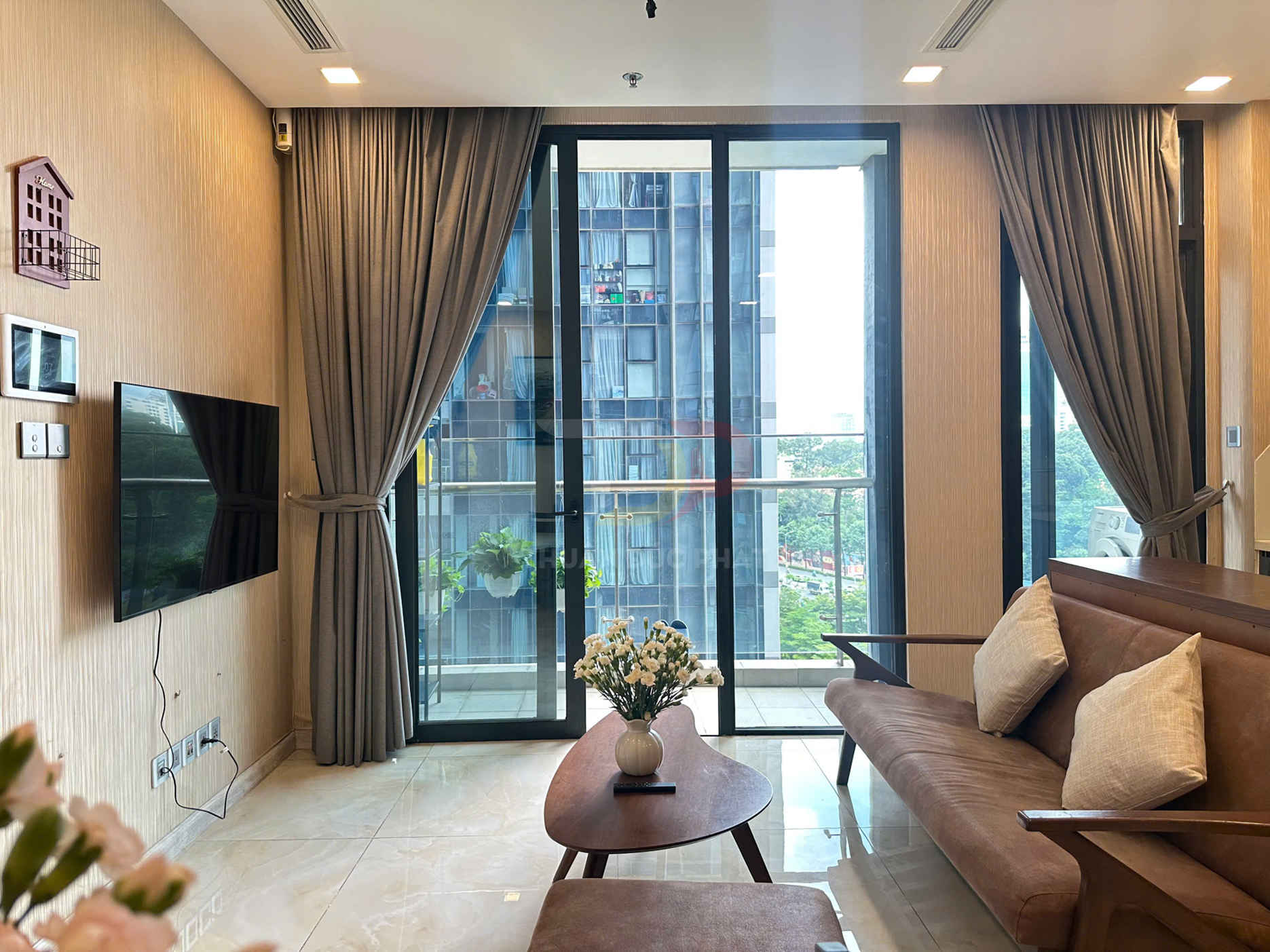 Phòng khách chung cư hiện đại với sofa nệm, bàn trà gỗ, tivi treo tường và cửa kính lớn nhìn ra ban công