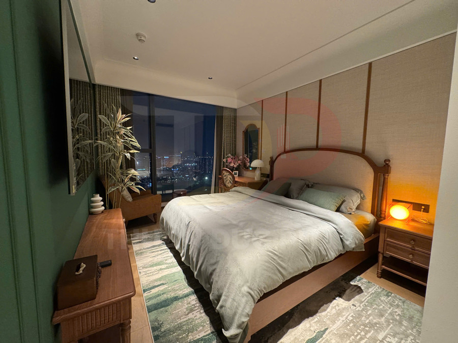 Phòng ngủ hiện đại với giường gỗ lớn, chăn ga màu xám xanh và đèn ngủ vàng ấm cạnh cửa sổ view thành phố