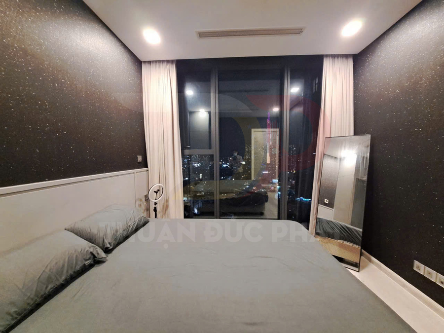 Phòng ngủ hiện đại với giường đôi ga xám, cửa kính lớn nhìn ra thành phố về đêm và gương đứng cạnh tường