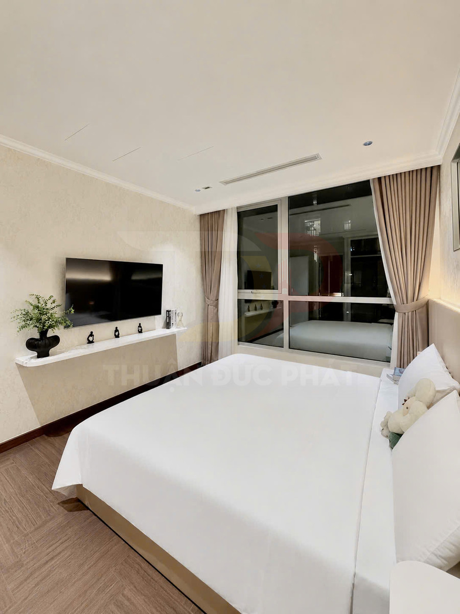 Phòng ngủ hiện đại với giường đôi ga trắng, tivi treo tường và cửa sổ lớn có rèm be