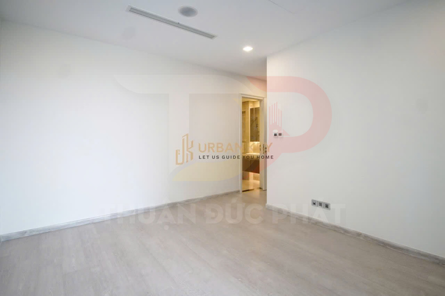 Phòng ngủ chung cư trống, tường trắng, sàn gỗ sáng màu, cửa dẫn vào phòng tắm có đèn downlight
