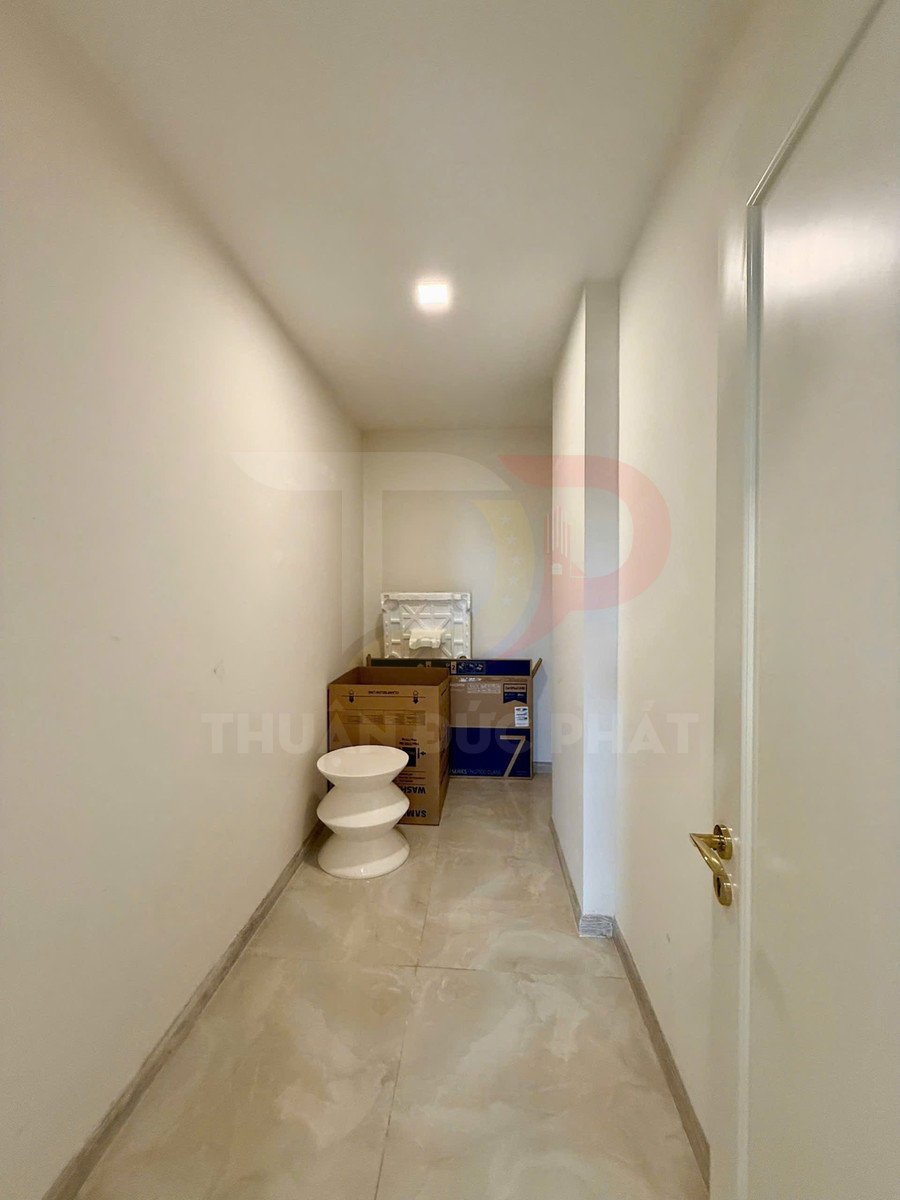 Hành lang nhỏ lát gạch sáng với ghế decor trắng và thùng carton đựng thiết bị điện tử cuối lối đi