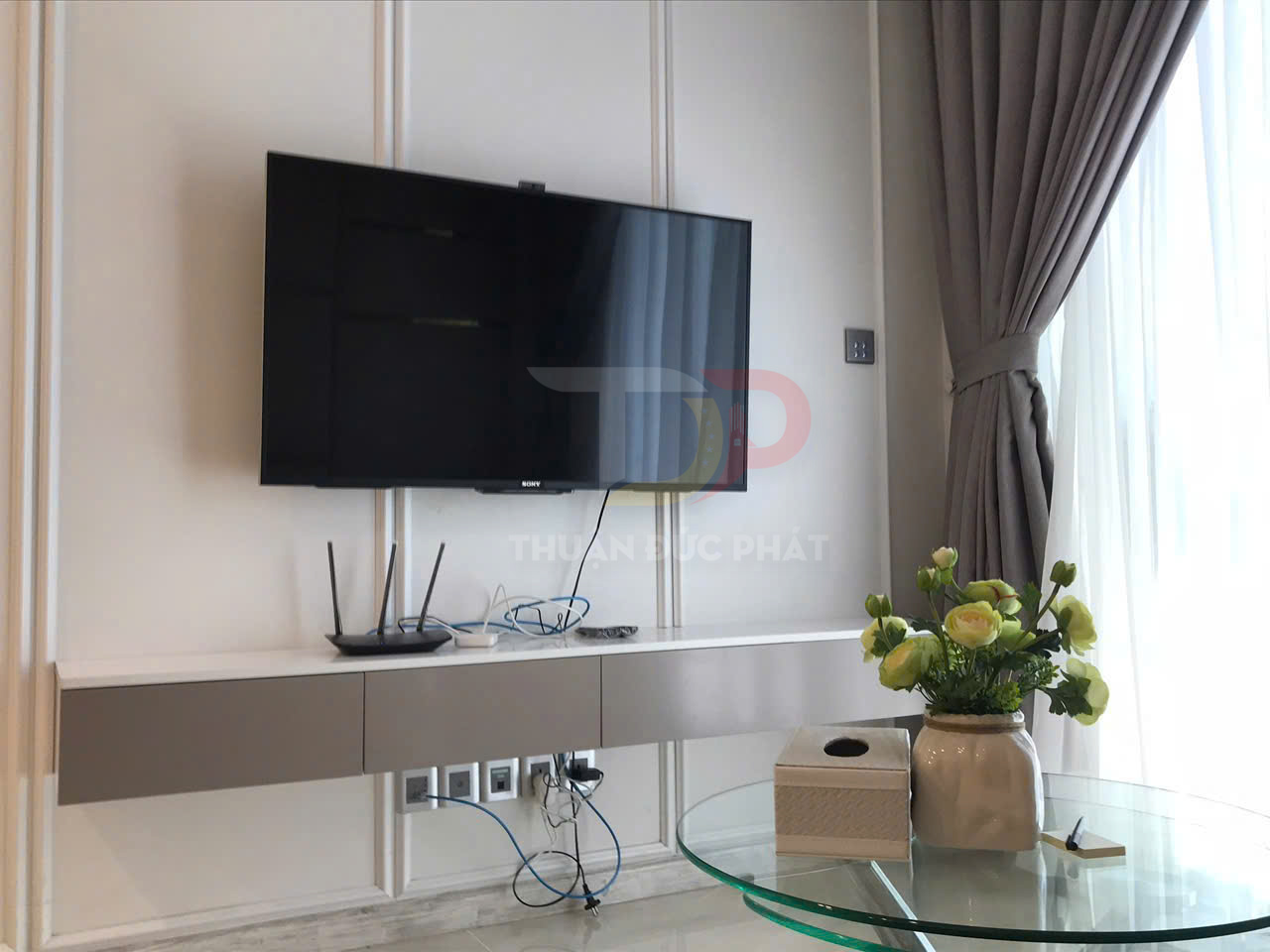 Tivi Sony treo tường với kệ treo hiện đại, bộ phát wifi và bàn kính đặt hộp khăn giấy cùng lọ hoa trang trí