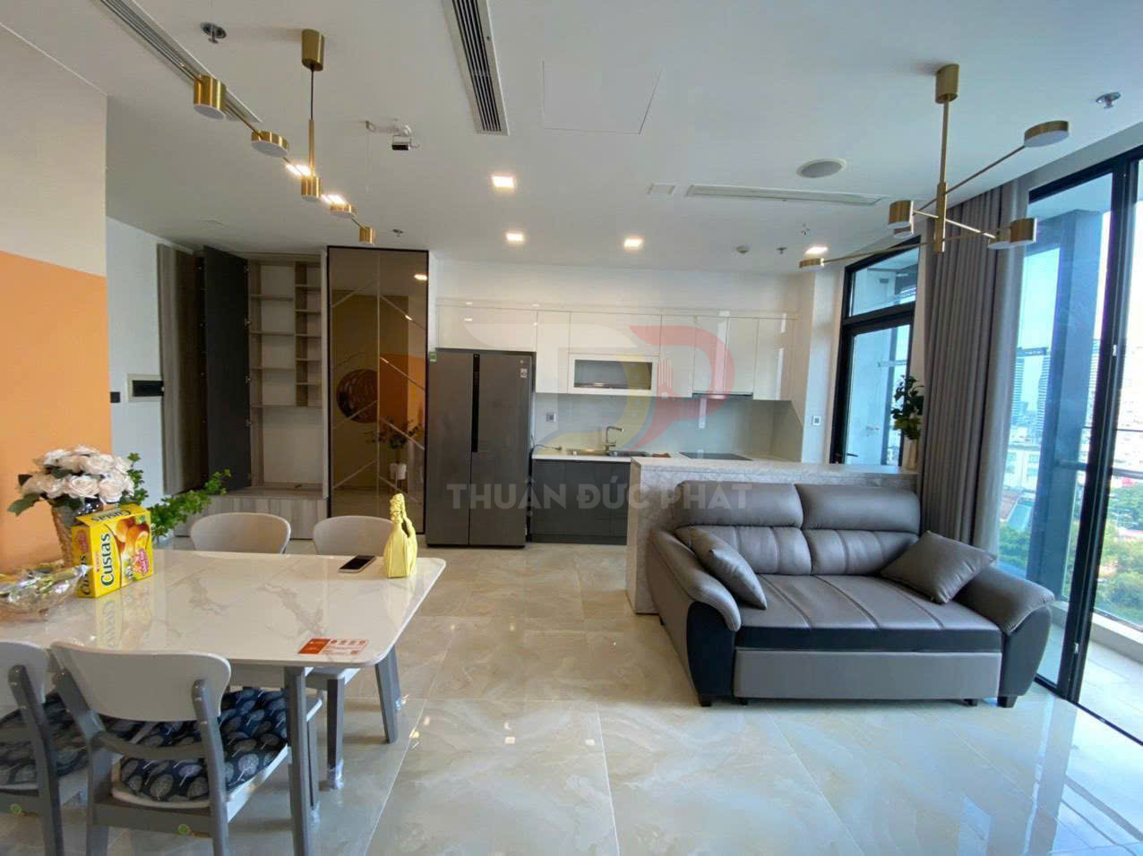 Phòng khách liền bếp hiện đại với sofa da xám, bàn ăn đá và tủ bếp trắng cạnh cửa kính lớn