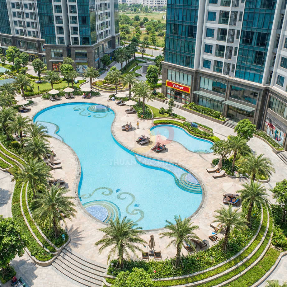 Hồ bơi ngoài trời hình uốn lượn giữa khu căn hộ cao tầng hiện đại, xung quanh là ghế tắm nắng và hàng dừa xanh