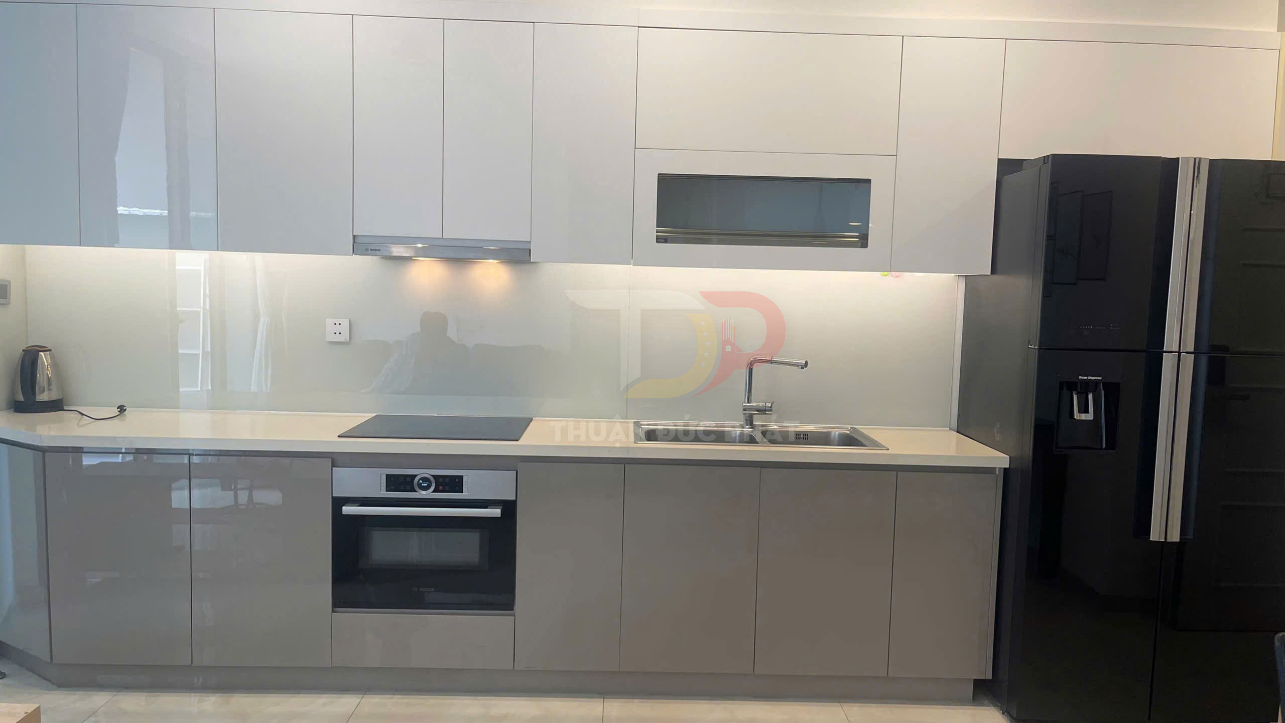 Tủ bếp hiện đại màu trắng xám với bếp từ, lò nướng Bosch, chậu rửa inox và tủ lạnh lớn màu đen