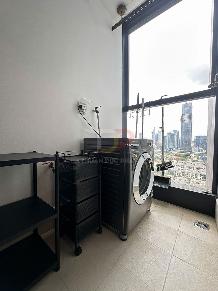 Máy giặt cửa trước màu bạc đặt cạnh kệ đựng đồ trong phòng giặt có cửa sổ lớn nhìn ra thành phố