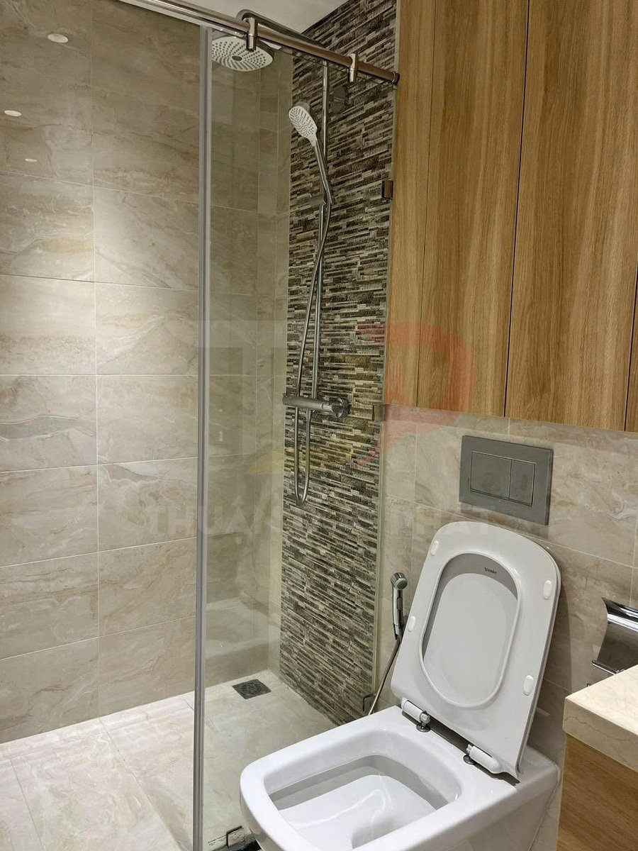 Bồn cầu hiện đại cạnh khu tắm kính với vòi sen đứng trong phòng tắm ốp gạch sáng màu