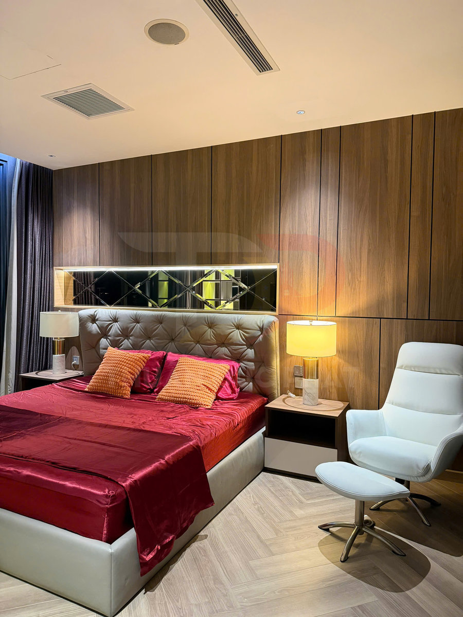 Giường ngủ bọc da với ga đỏ, gối cam, kèm ghế thư giãn trắng và đèn bàn trong phòng ngủ hiện đại