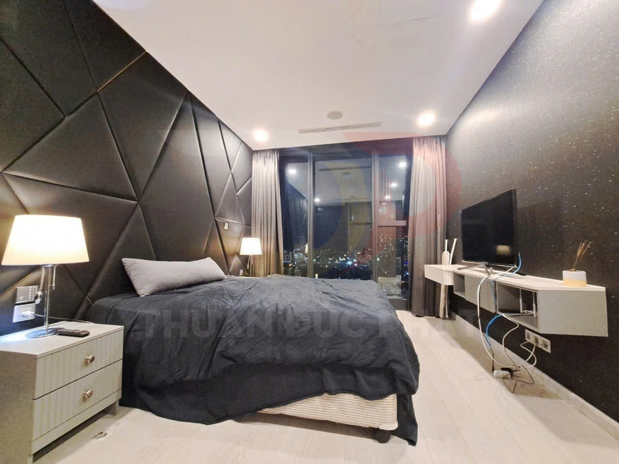 Phòng ngủ hiện đại với giường đôi chăn màu xám đậm, tường ốp da đen và tivi treo tường đối diện
