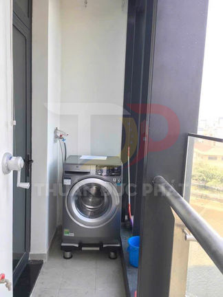 Máy giặt cửa trước đặt ở ban công chung cư, nối với vòi nước, cạnh xô nhựa xanh