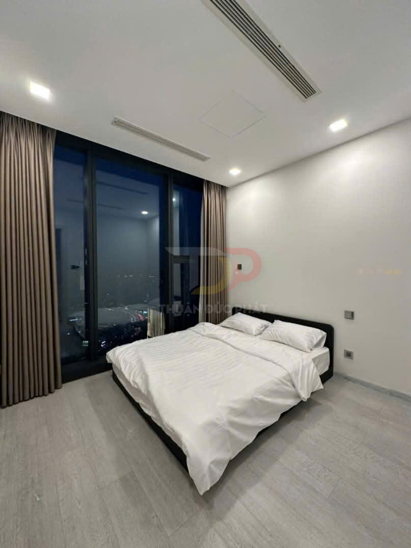 Phòng ngủ tối giản với giường đôi bọc nệm trắng cạnh cửa sổ kính lớn nhìn ra thành phố ban đêm