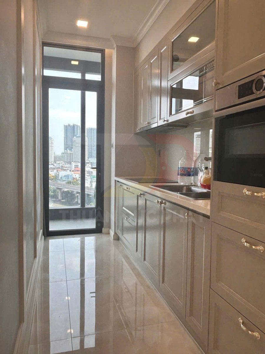 Căn bếp chung cư nhỏ với tủ bếp gỗ màu kem, mặt đá, bồn rửa inox và cửa kính lớn nhìn ra thành phố