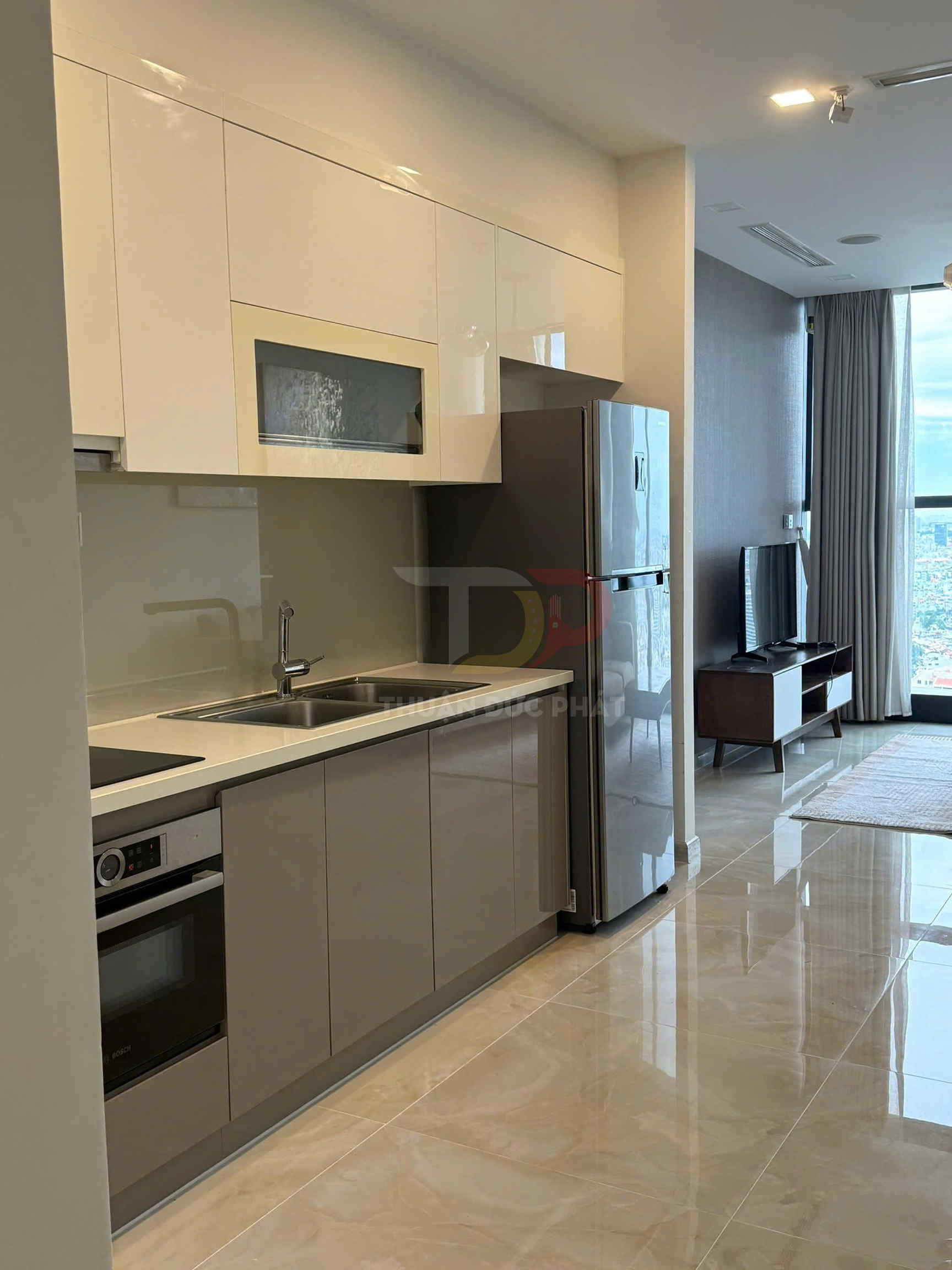 Gian bếp chung cư hiện đại với tủ bếp chữ I, bếp điện, lò nướng âm tủ và tủ lạnh inox cạnh phòng khách