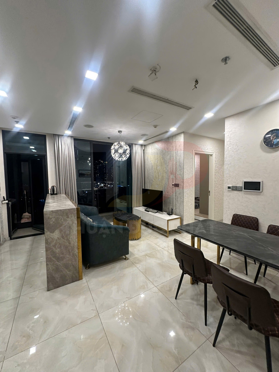 Phòng khách chung cư hiện đại với sofa, bàn ăn đá, kệ tivi và cửa kính lớn nhìn ra ban công ban đêm