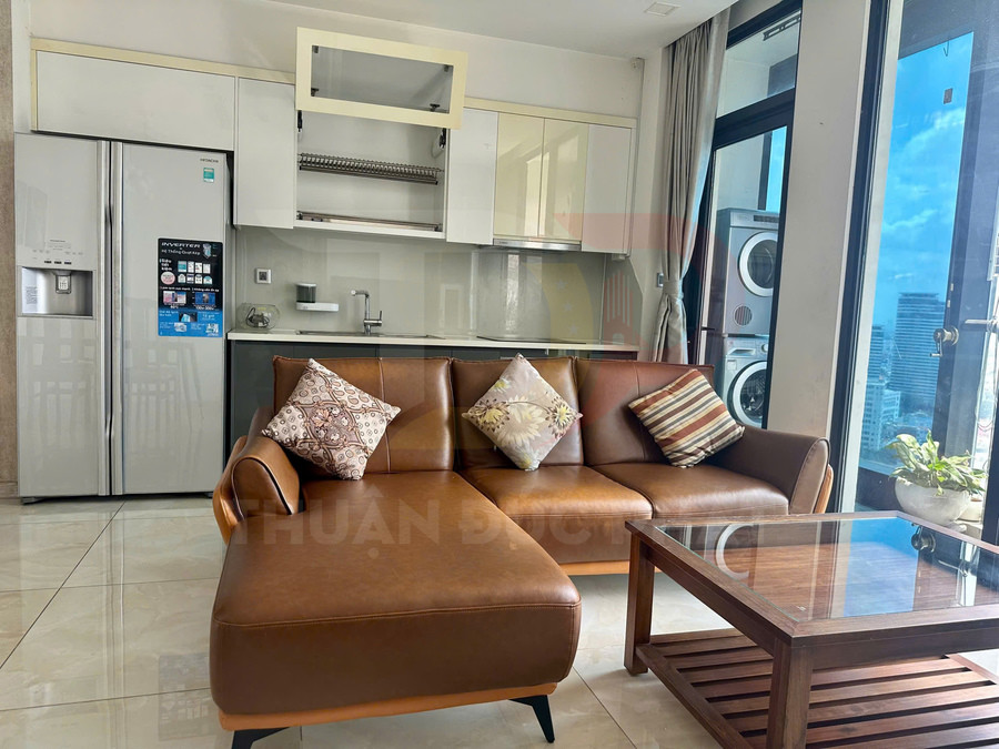 Sofa da chữ L màu nâu với bàn trà gỗ trong phòng khách liền bếp hiện đại có tủ lạnh và cửa kính lớn