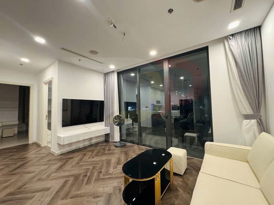 Phòng khách chung cư hiện đại với sofa kem, bàn trà mặt kính đen khung vàng và tivi treo tường cạnh cửa kính lớn