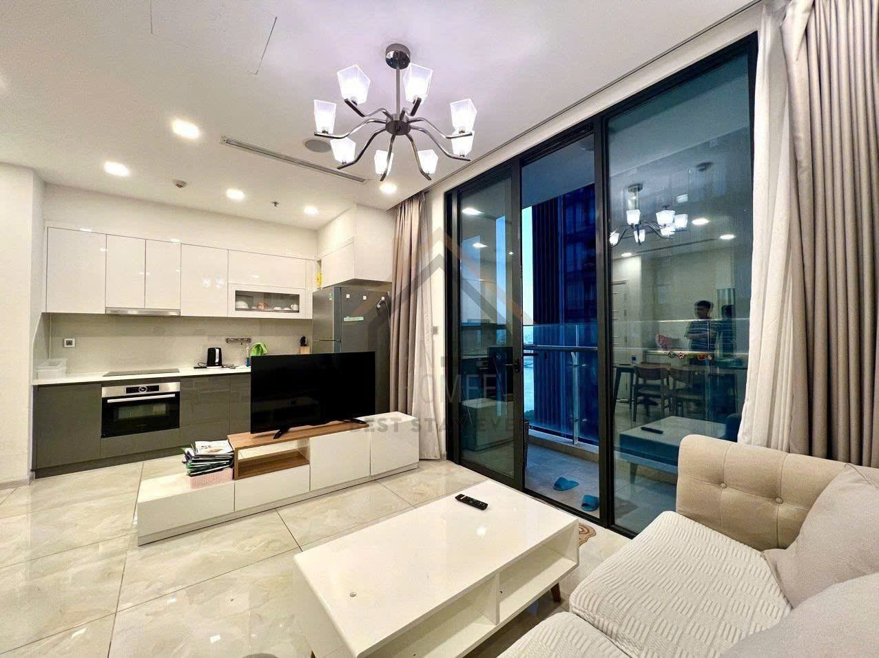 Phòng khách căn hộ hiện đại với bếp liền kề, sofa sáng màu, bàn trà và tivi trên kệ trắng cạnh cửa kính lớn