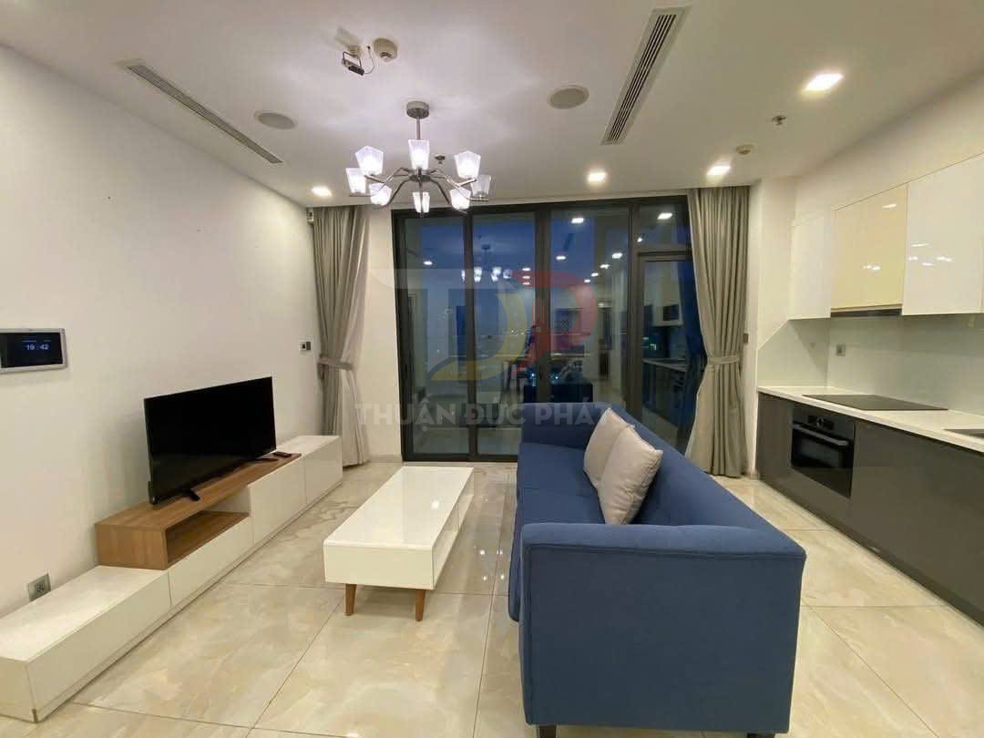 Phòng khách căn hộ hiện đại với sofa xanh, bàn trà trắng, tivi màn phẳng và bếp liền kề