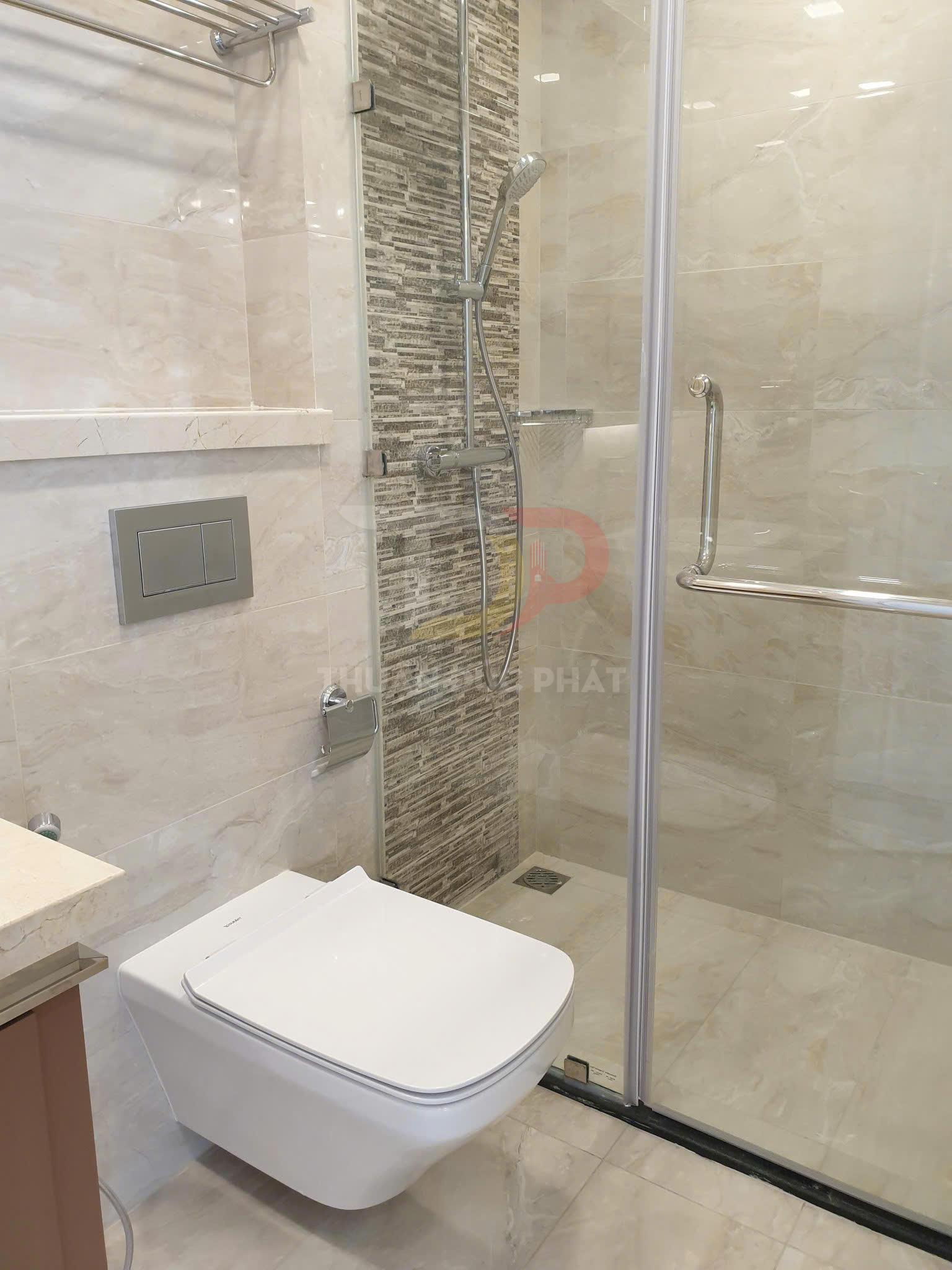 Bồn cầu treo tường hiện đại cạnh buồng tắm kính và vòi sen trong phòng tắm lát đá sáng