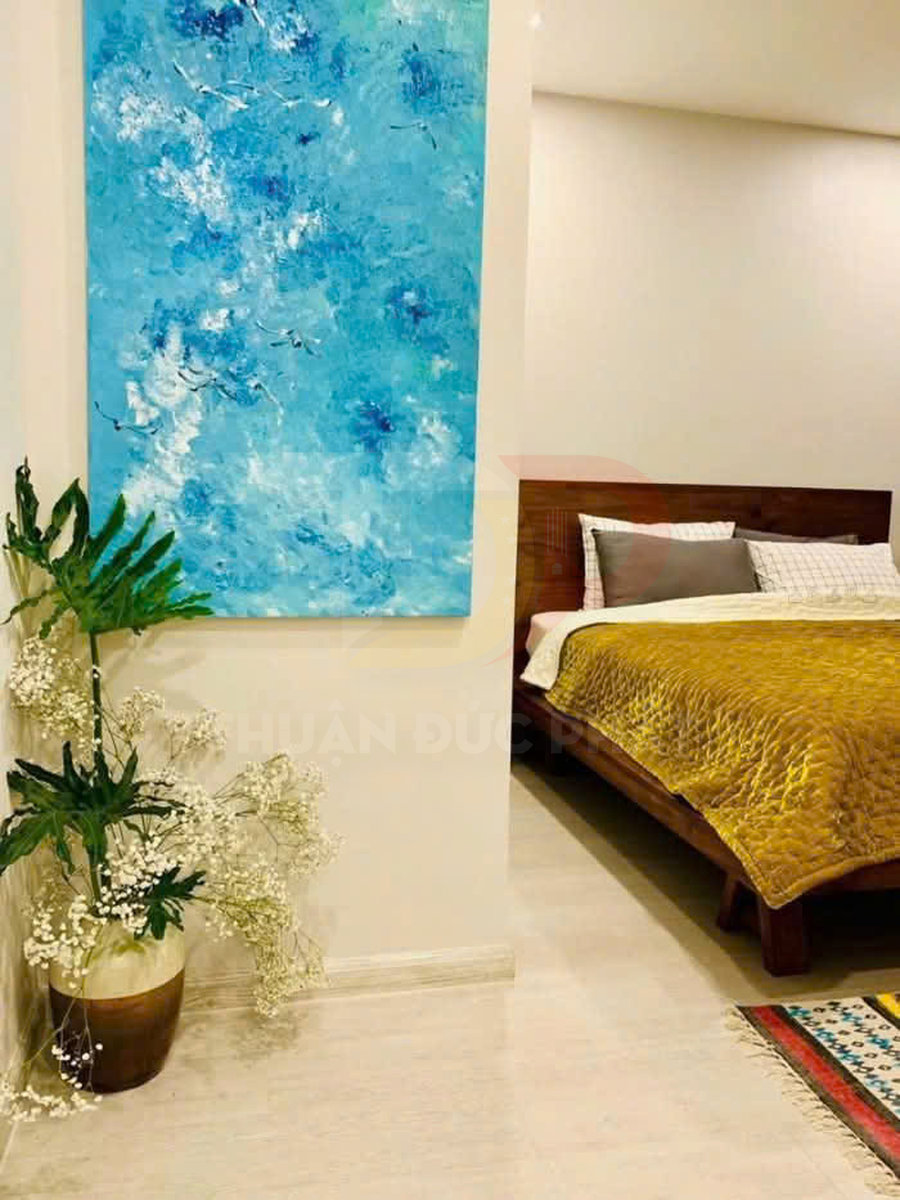 Tranh trừu tượng màu xanh treo tường cạnh chậu cây trang trí trong phòng ngủ giường gỗ chăn vàng