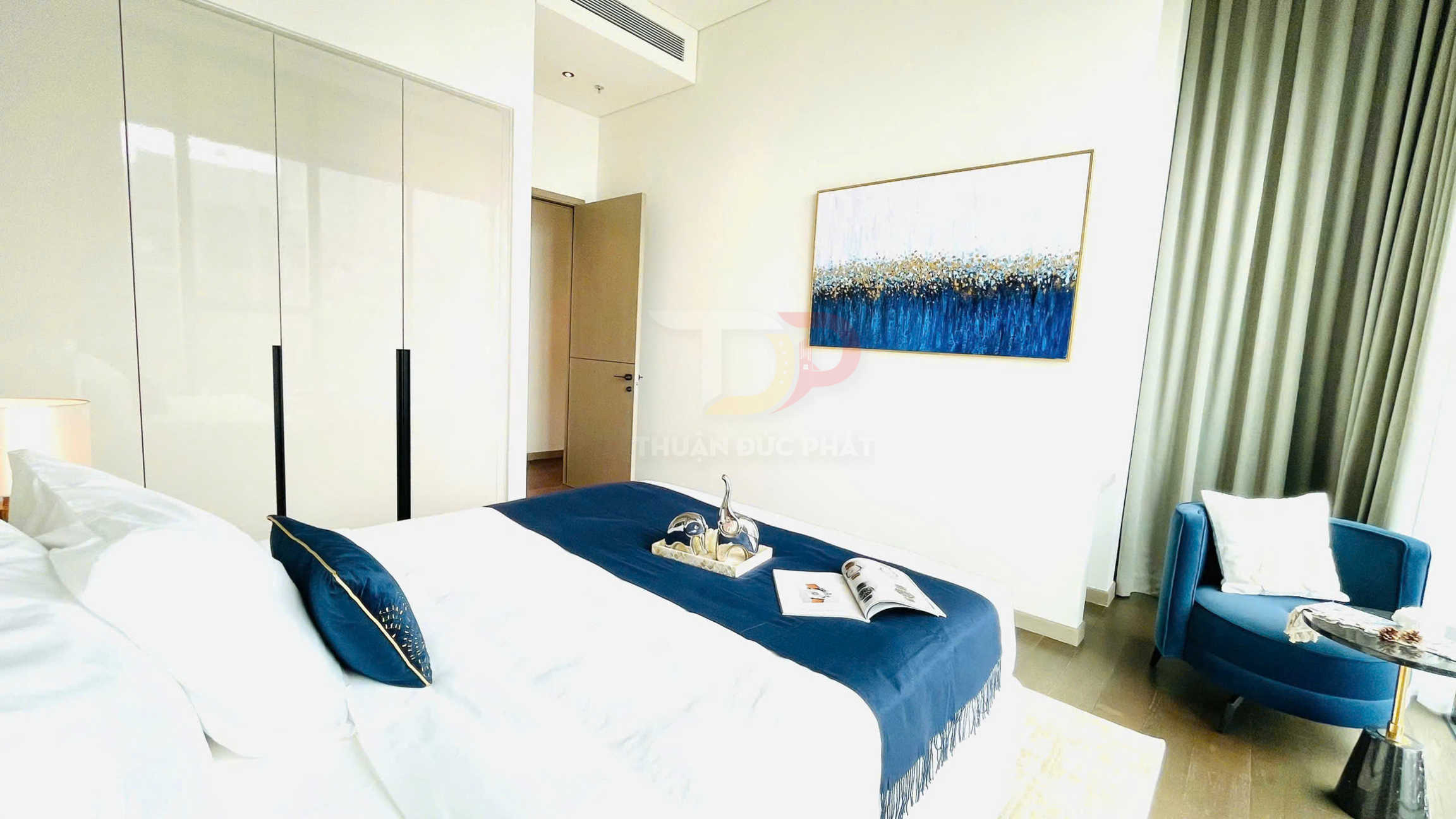 Phòng ngủ hiện đại với giường trải ga trắng chăn xanh, tủ áo cửa lùa và ghế bành xanh cạnh bàn trà