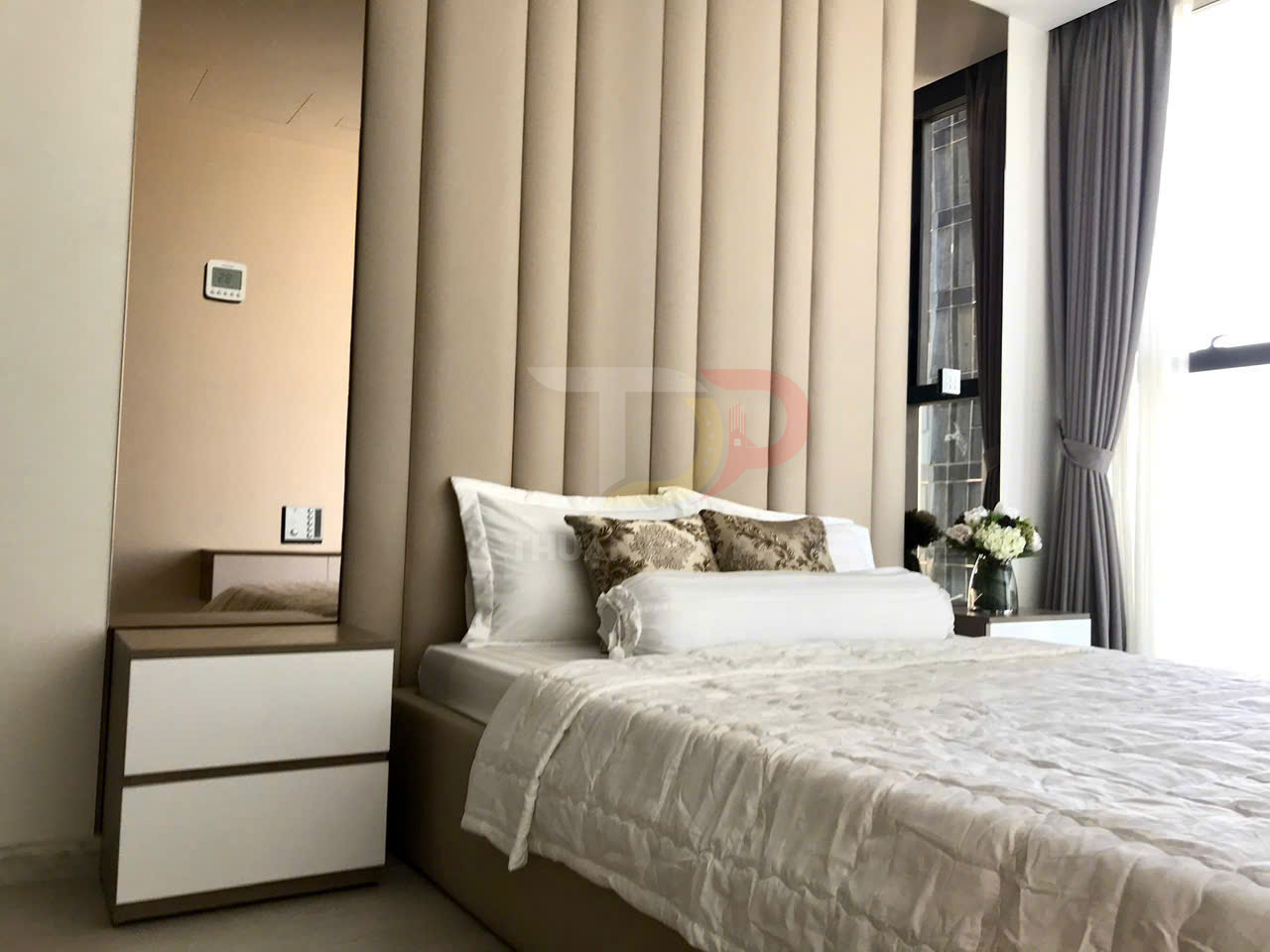 Phòng ngủ hiện đại với giường bọc nệm màu be, ga gối trắng và tủ đầu giường cạnh cửa sổ có rèm xám