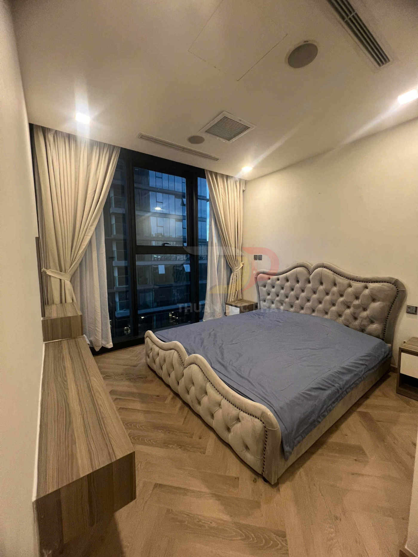 Phòng ngủ hiện đại với giường bọc nệm màu be, ga xanh, sàn gỗ và cửa sổ lớn rèm trắng