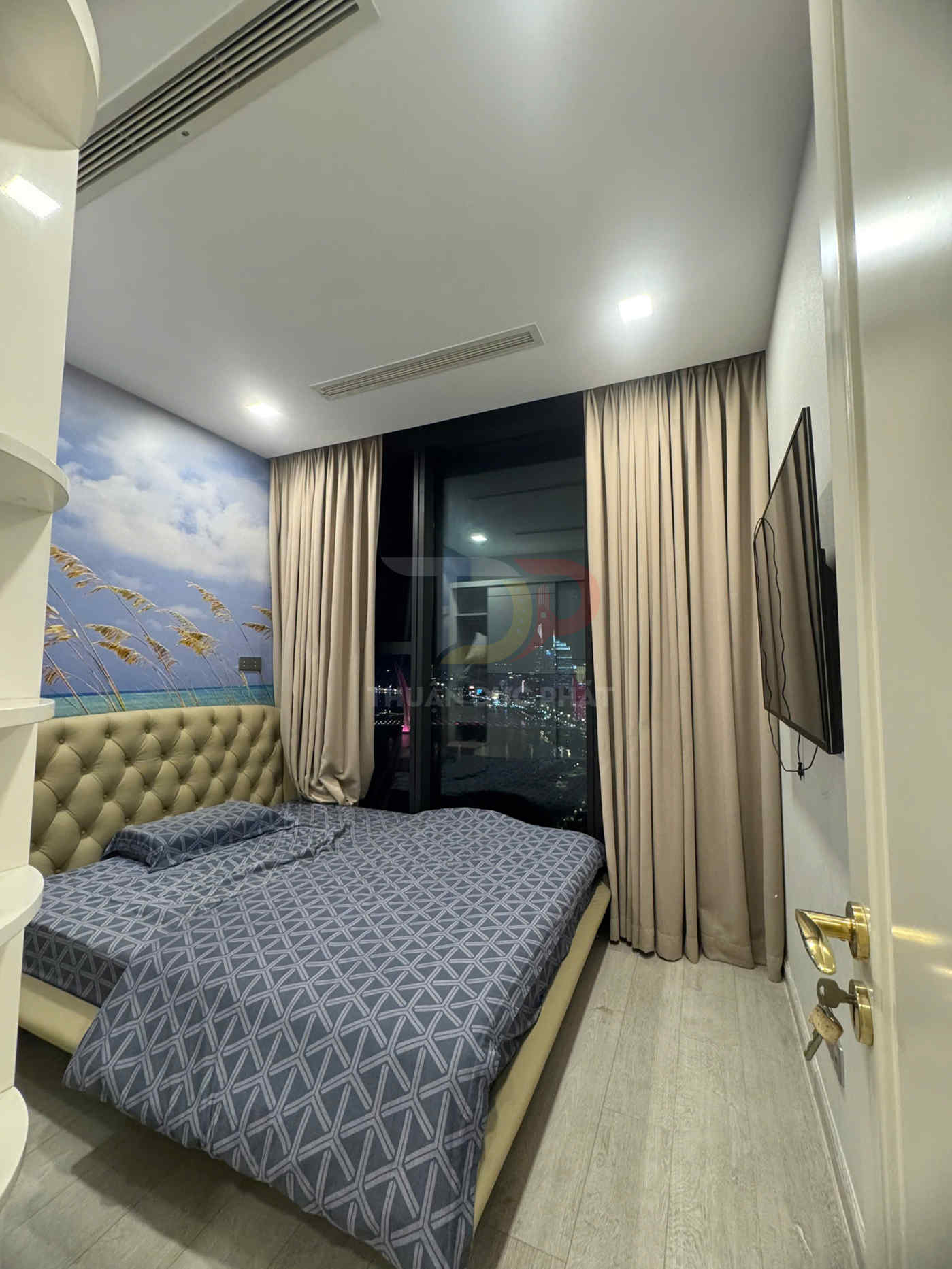 Phòng ngủ nhỏ với giường đôi bọc da, chăn ga họa tiết xám và cửa sổ kính lớn nhìn ra thành phố ban đêm