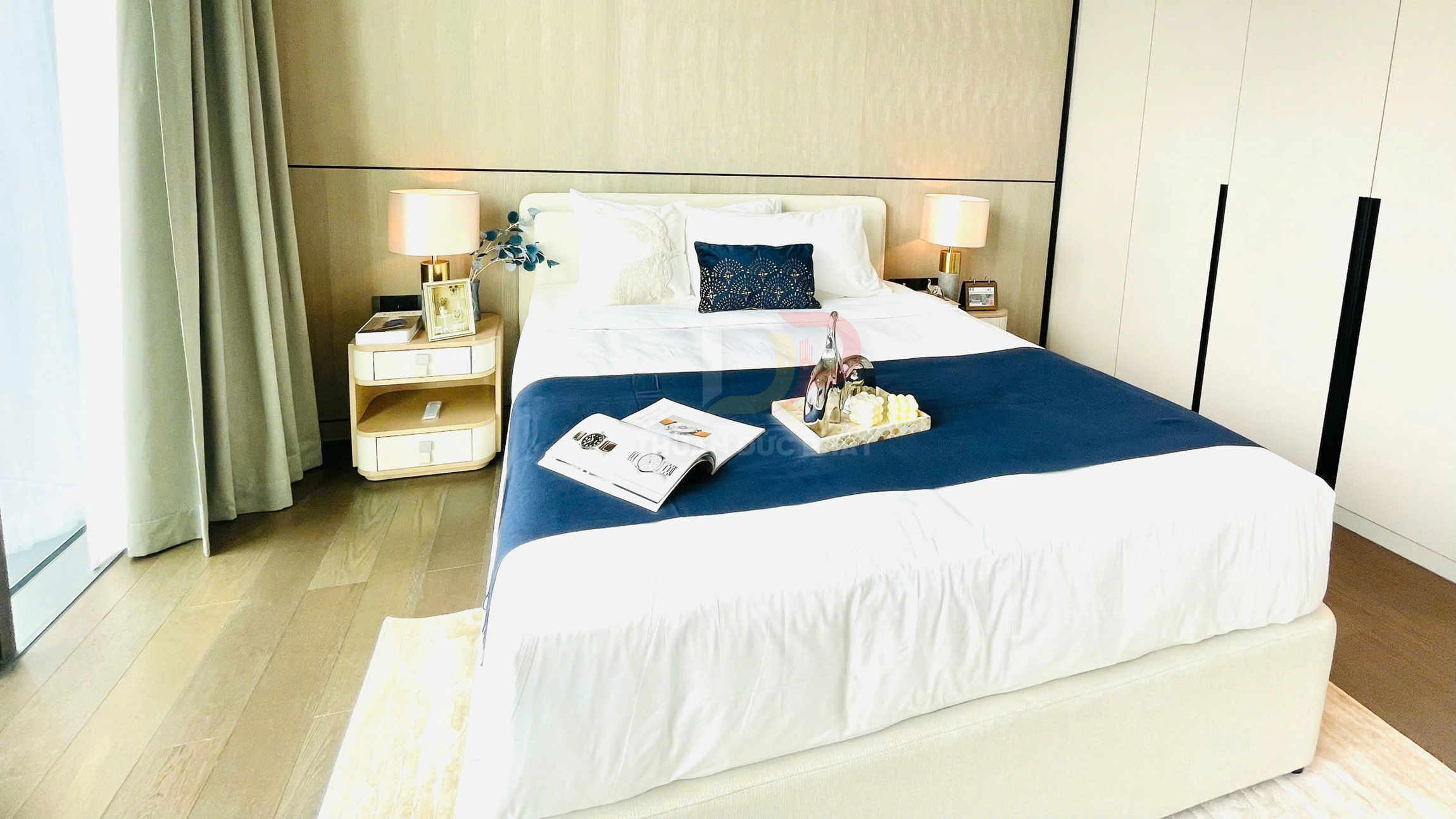 Giường ngủ hiện đại trải ga trắng chăn xanh, có khay trang trí và tạp chí đặt trên nệm trong phòng ngủ sáng