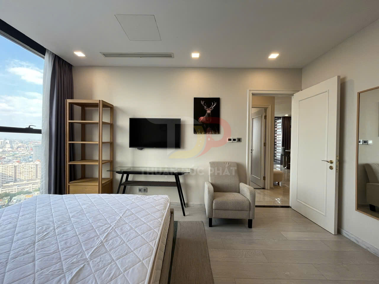 Phòng ngủ chung cư hiện đại với giường nệm trắng, ghế bành vải be, kệ gỗ và tivi treo tường cạnh cửa sổ lớn