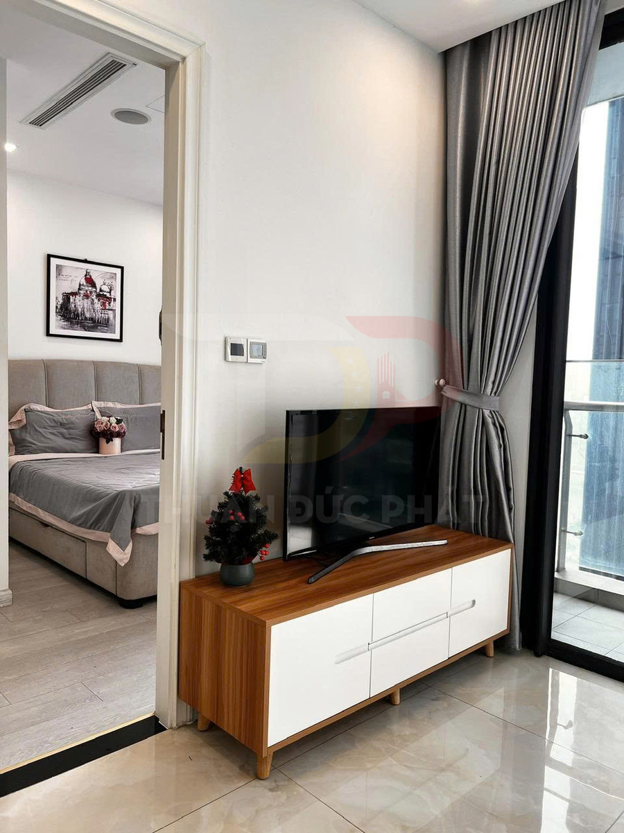 Kệ tivi gỗ hiện đại đặt cạnh cửa sổ, phía sau là phòng ngủ giường bọc nệm màu xám