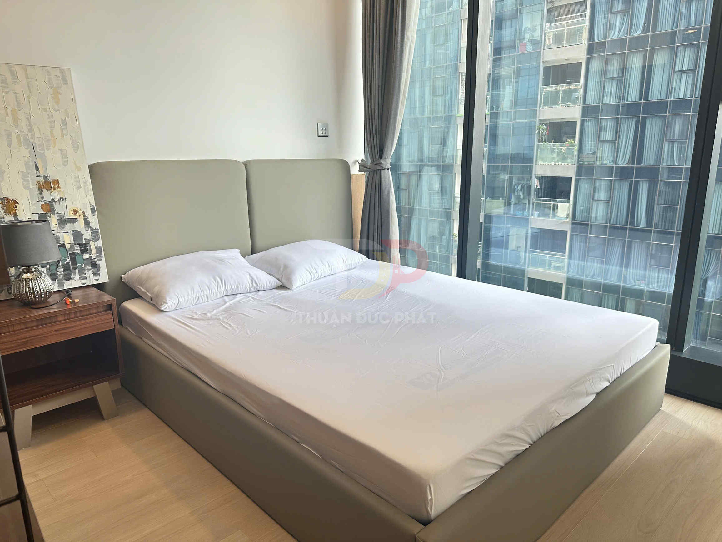 Giường ngủ bọc da màu xám với nệm trắng và hai gối, đặt cạnh cửa kính lớn nhìn ra chung cư